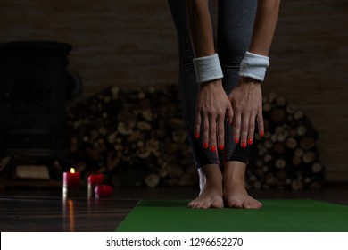 Young Lady Standing On The Yoga Mat And Bending Down While Trying To Touch The Toes. Firewood And Candles On The Background