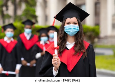 Young Lady Student In Graduation Costume And Protective Face Mask Showing Her Diploma, Female Student Posing Over International Group Of Students At University Campus, Copy Space