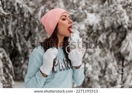 A young lady in a sweater, hat and knitted mittens in a snowy forest holds a mug of snow in her hands and licks it off. Cheerful winter mood.