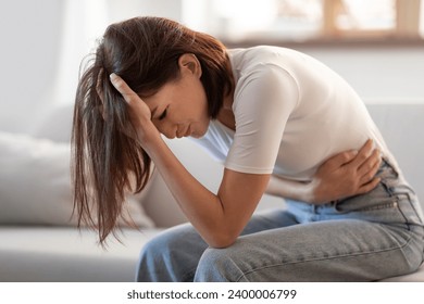 Young lady touching her stomach and head in distress, suffering from headache and stomachache, having symptoms of PMS and gastrointestinal discomfort, sitting in living room at home - Powered by Shutterstock