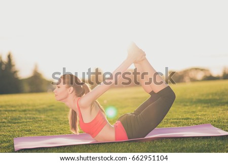 Young lady trainer is making stretching lying outdoors in spring park on a purple mat on nice green grass, so fit and healthy, wearing modern pink and black sport outfit, with tail