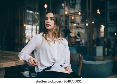 Young lady in trendy clothes sitting near wooden table and flipping pages in notebook while resting in contemporary cafe and looking dreamily away