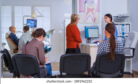 Young Lady Visiting Stomatological Clinic For Teeth Checking While Dentistry Doctor Preparing Old Man For Dental Surgery In Background. Patients Sitting In Crowded Waiting Room Of Orthodontist Office