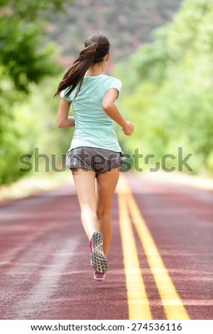 Young lady working out running away on rural road. Woman runner athlete training jogging during workout outside. Full body length rear view showing back. Girl in shorts and running shoes.