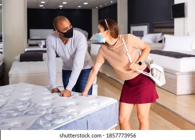 Young Latin American Couple In Protective Masks Choosing New Comfortable Mattress Together At Furniture Shop. Concept Of Shopping Precautions In Pandemic