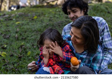 Young Latin American Family Sitting On The Grass Spending The Afternoon In The Park And Playing With Their 3 Year Old Child. Family Concept.