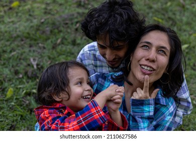 Young Latin American Family Sitting On The Grass Spending The Afternoon In The Park And Playing With Their 3 Year Old Child. Family Concept.