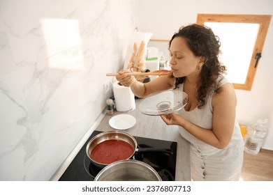 Young Latin American pretty housewife tasting the tomato sauce she's cooking in the home kitchen. People. Culinary. Lifestyle - Powered by Shutterstock