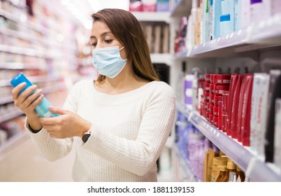 Young Latin American Woman In Protective Face Mask Carefully Choosing Hair Care Products At Cosmetics Shop. Pandemic Shopping Precautions 