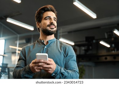 Young Latin business man entrepreneur holding mobile phone standing in office. Confident professional businessman investor executive using cellphone at work looking away thinking of digital ideas. - Powered by Shutterstock