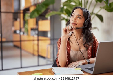 Young latin business woman wearing headphones with microphone daydreaming while working on laptop. Thoughtful mixed race businesswoman take a break and thinking about new idea. 