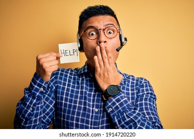 Young latin call center agent man overworked holding help message paper using headset cover mouth with hand shocked with shame for mistake, expression of fear, scared in silence, secret concept