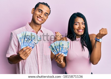 Young latin couple holding south african rand banknotes screaming proud, celebrating victory and success very excited with raised arm 