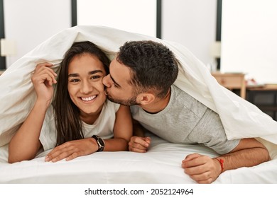 Young Latin Couple Kissing And Covering With Bed Sheet Lying On The Bed.