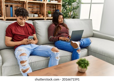 Young Latin Couple With Serious Expression Using Laptop And Smartphone At Home.