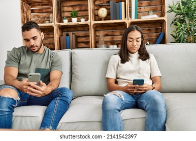 Young Latin Couple With Serious Expression Using Smartphone At Home.