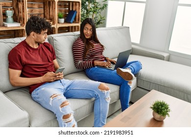 Young Latin Couple With Serious Expression Using Laptop And Smartphone At Home.