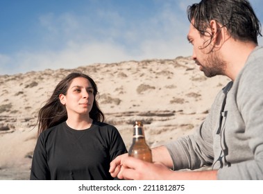 Young Latin Couple Sitting On The Beach Talking And Drinking