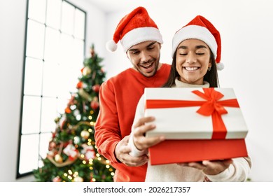 Young Latin Couple Smiling Happy And Hugging Unboxing Christmas Gift At Home.