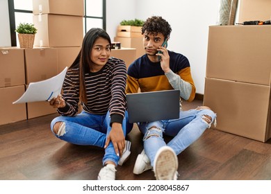 Young Latin Couple Using Laptop And Talking On The Smartphone Sitting On The Floor At New Home.