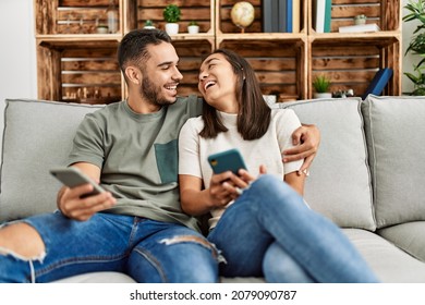 Young Latin Couple Using Smartphone Sitting On The Sofa At Home.