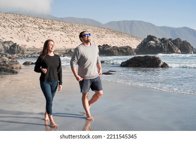 Young Latin Couple Walking Along The Shore Of The Beach Barefoot Happy