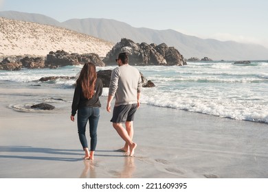 Young Latin Couple Walking Along The Shore Of The Beach Barefoot  Back View