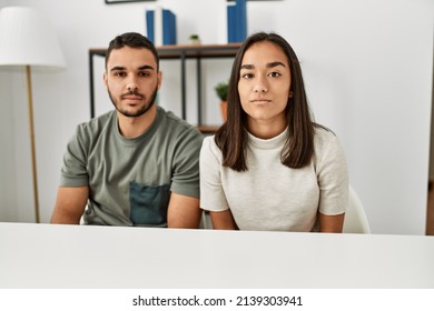 Young Latin Couple Wearing Casual Clothes Sitting On The Table With Serious Expression On Face. Simple And Natural Looking At The Camera. 