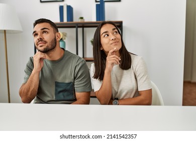 Young Latin Couple Wearing Casual Clothes Sitting On The Table Serious Face Thinking About Question With Hand On Chin, Thoughtful About Confusing Idea 