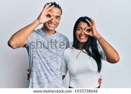 Young latin couple wearing casual clothes doing ok gesture with hand smiling, eye looking through fingers with happy face. 