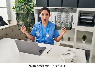 Young Latin Doctor Woman Holding X-ray In Shock Face, Looking Skeptical And Sarcastic, Surprised With Open Mouth 