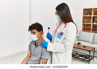 Young Latin Doctor Woman Wearing Medical Mask Auscultating The Ear Of Man Using Diapason At Examination Room.