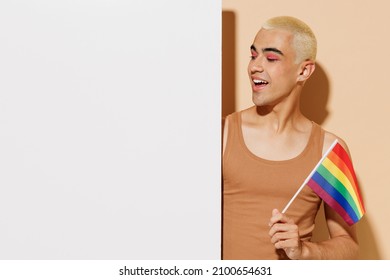 Young Latin Gay Man In Beige Tank Shirt Holding Big White Empty Blank Poster Billboard For Promotional Content, Place For Text Or Image Rainbow Flag Isolated On Plain Light Ocher Background Studio.