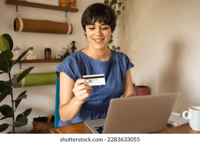 Niña latina joven con el pelo corto sonriendo mientras sostiene una tarjeta de crédito. Está sentada en un apartamento lleno de plantas verdes y usando una laptop.