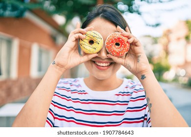 Young latin girl smiling happy holding donuts over eyes walking at the city.
