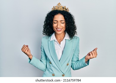 Young latin girl wearing business clothes and queen crown very happy and excited doing winner gesture with arms raised, smiling and screaming for success. celebration concept. 