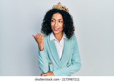 Young Latin Girl Wearing Business Clothes And Queen Crown Smiling With Happy Face Looking And Pointing To The Side With Thumb Up. 