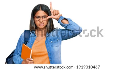 Young latin girl wearing student backpack and holding books making fun of people with fingers on forehead doing loser gesture mocking and insulting. 