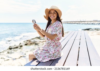 Young latin girl wearing summer using smartphone sitting on the bench hat at the beach. - Powered by Shutterstock