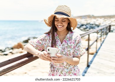Young latin girl wearing summer hat using smartphone at the beach. - Shutterstock ID 2226621703
