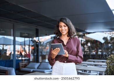 Young latin Hispanic adult professional business woman looking at online trade app. European mature businesswoman CEO holding digital tablet using fintech tab application working in workspace office