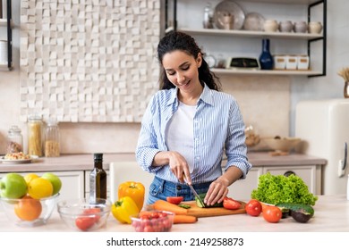 Young latin lady cooking delicious and healthy salad standing in modern kitchen, housewife maid preparing vegetable for lunch, slicing cucumber with knife on cutting board - Powered by Shutterstock