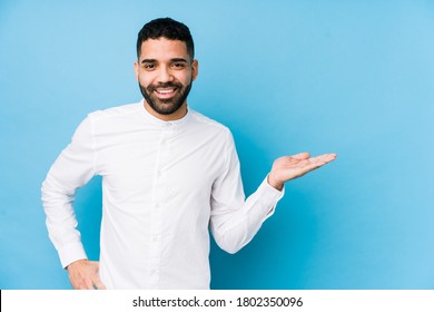 Young Latin Man Against A Blue  Background Isolated Showing A Copy Space On A Palm And Holding Another Hand On Waist.