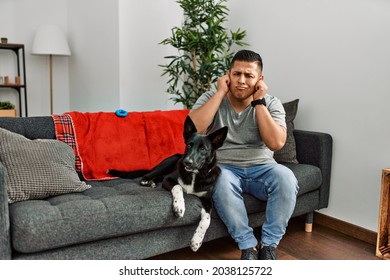 Young Latin Man And Dog Sitting On The Sofa At Home Covering Ears With Fingers With Annoyed Expression For The Noise Of Loud Music. Deaf Concept. 