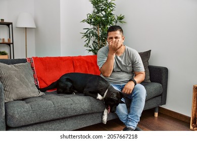 Young latin man and dog sitting on the sofa at home smelling something stinky and disgusting, intolerable smell, holding breath with fingers on nose. bad smell 