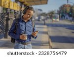 Young latin man with headphones looking at his mobile phone at the bus stop.