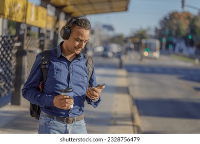 Young latin man with headphones looking at his mobile phone at the bus stop. - Powered by Shutterstock