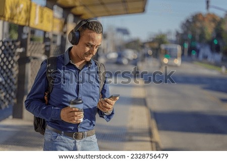 Young latin man with headphones looking at his mobile phone at the bus stop.