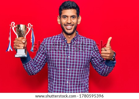 Young latin man holding winner trophy smiling happy and positive, thumb up doing excellent and approval sign 