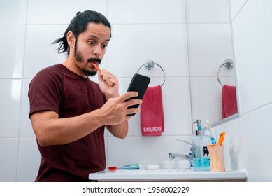 Young Latin Man With Long Black Hair Using The Phone While Brushing Teeth At The Bathroom